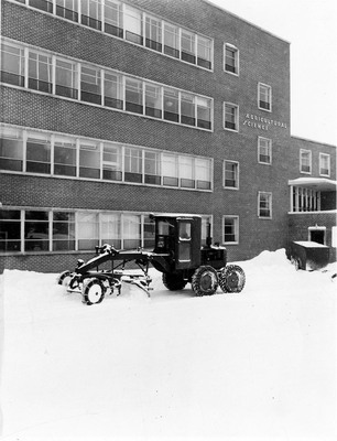 A picture of farming equipment equipped with a snow plow outside of the Agricultural Science Building. Snow can be seen on the ground.