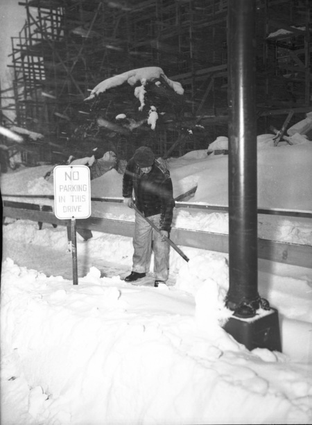 A man shoveling snow next to a sign that reads 'No Parking in this Drive'. A building under construction can be seen in the background.