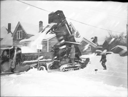 A snow excavator pouring snow into a dump truck during a snowstorm, three men can be seen operating the truck and excavator.