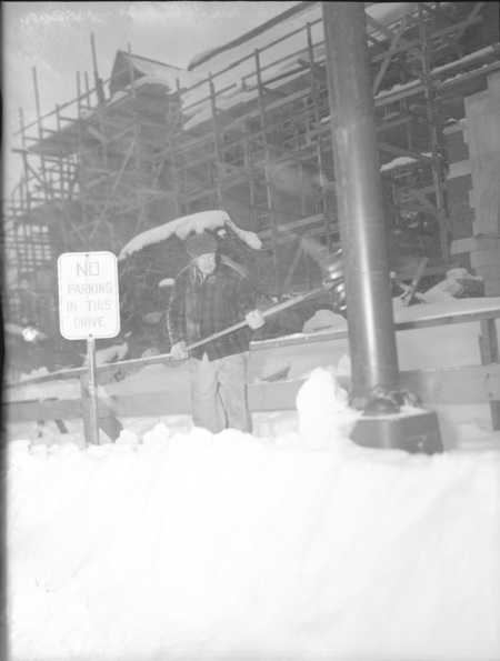 A man shoveling snow next to a sign that reads 'No Parking in this Drive'. A building under construction can be seen in the background.