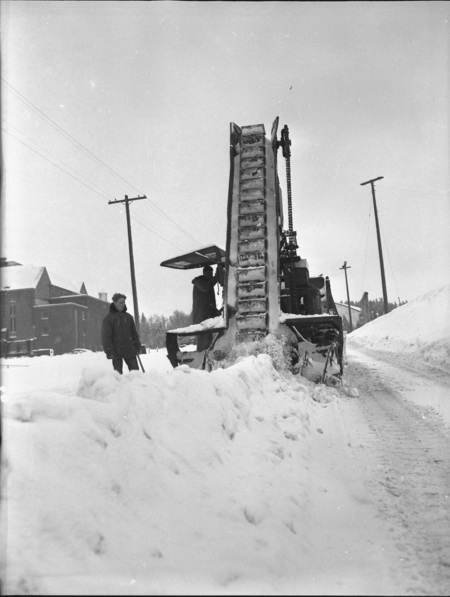 A snow excavator pouring snow into a dump truck during a snowstorm, three men can be seen operating the truck and excavator.