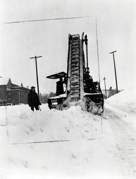 A snow excavator pouring snow into a dump truck during a snowstorm, three men can be seen operating the truck and excavator.