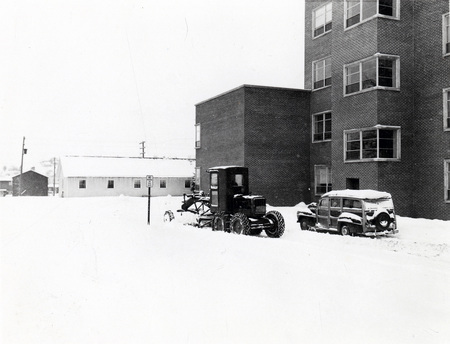A picture of farming equipment equipped with a snow plow and a automobile outside of the Agricultural Science Building during winter.