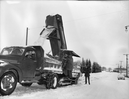 A snow excavator pouring snow into a dump truck during a snowstorm, three men can be seen operating the truck and excavator.