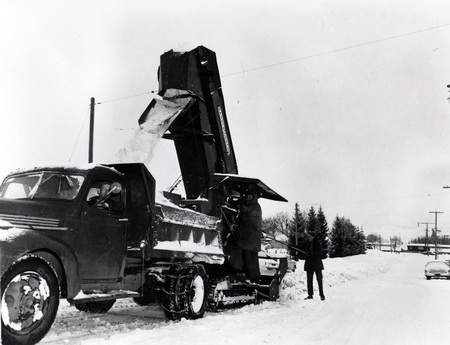 A snow excavator pouring snow into a dump truck during a snowstorm, three men can be seen operating the truck and excavator.