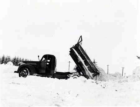 A dump truck dumping snow, the driver can be seen with the truck door open.