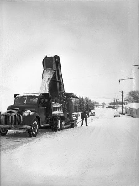 A snow excavator pouring snow into a dump truck during a snowstorm, three men can be seen operating the truck and excavator.