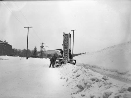 A snow excavator pouring snow into a dump truck during a snowstorm, three men can be seen operating the truck and excavator.