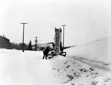 A snow excavator pouring snow into a dump truck during a snowstorm, three men can be seen operating the truck and excavator.