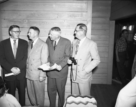 President J.E. Buchanan (right)  holds a pair of binoculars and stands near three other men. There are other people in the background.