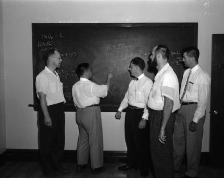Dr. K. A. Bush writing on a chalkboard while visiting mathematicians from various fields.