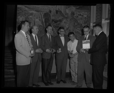 A group photo of bowling champions holding their awards in the Agricultural Science building with College of Agriculture Dean James E. Kraus. The Kirkwood Mural is seen behind them.