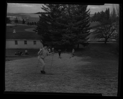 Students in ski class practice going downhill on skis without snow in a grassy hill behind the Radio-TV Building just south of the Administration Building. Memorial Gym and MacLean Field can be seen in the background.