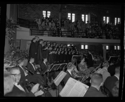 The Vandaleers, led by Professor Glen Lockery, sing "Here We Have Idaho" while the University Symphony Orchestra looks on at commencement in Memorial Gym.