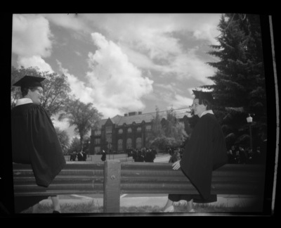 Two graduating senior women sitting in their caps and gowns on a bench during Commencement outside the Administration Building.