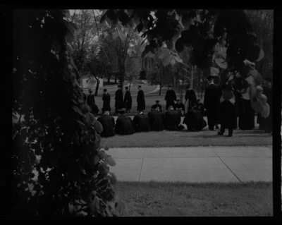 Graduates in cap and gowns rest on the grass outside of the Administration Building while waiting to file into Memorial Gym for Commencement.