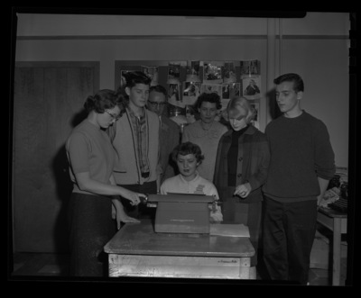 Students stand around Delores Hanson at her type writer during a journalism conference at the University of Idaho. Dr. Granville Price, professor of journalism, and Mrs. Stuart observe from behind.