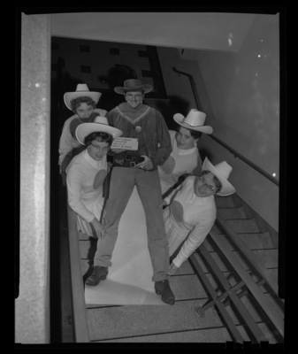 A photograph taken from above in a SUB stairwell of four members of the Spurs (l-r) Sharon Lance, Linda Smith, Katherine Koelsch, and Blanche Blecha carrying a cut out of Gunsmoke Star, James Arness that was used during the Idaho Cattlemen's Association meeting.