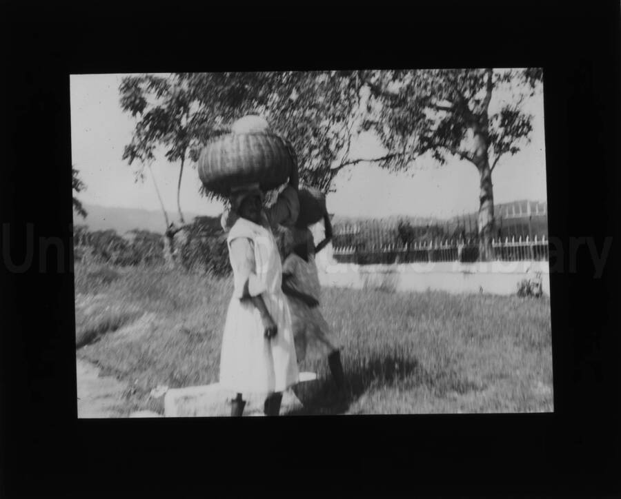 A woman and a girl carrying baskets on their heads. An iron gate can be seen in the background.