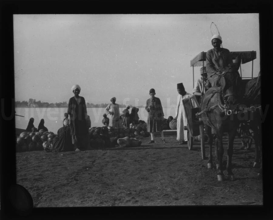 Maud Iddings poses on a shore, alongside other persons and a four wheeled carriage pulled by horses.
