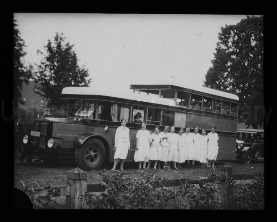 Ten women dressed in white stand on the side of a bus, with passengers looking through the windows.