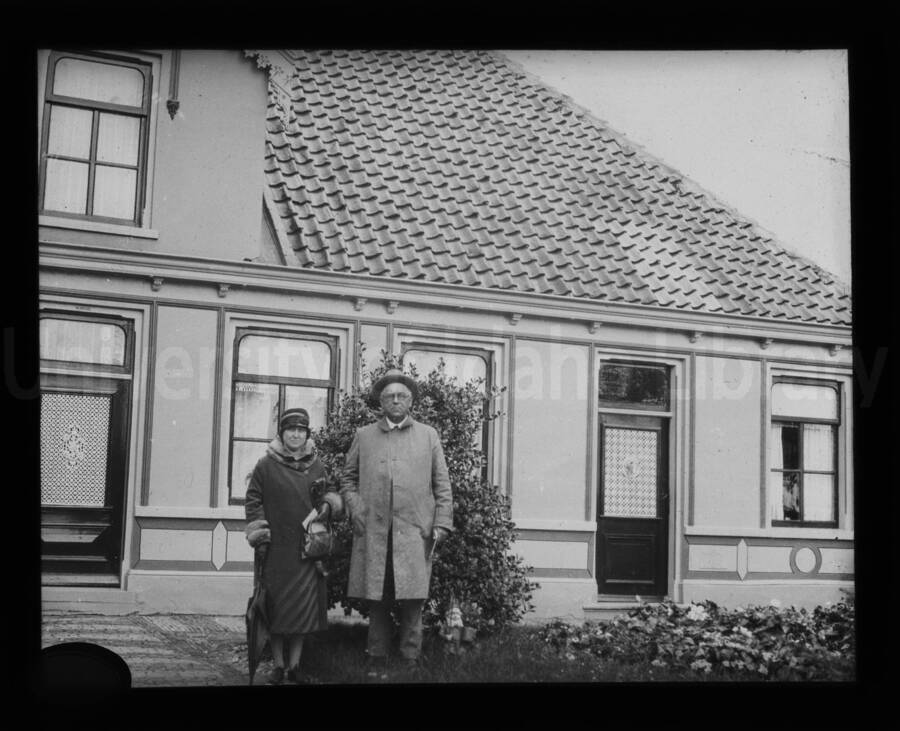 A woman and a man in front of a house with terracotta tile shingles on the roof. A child can be seen looking out a window on the far right of the image.