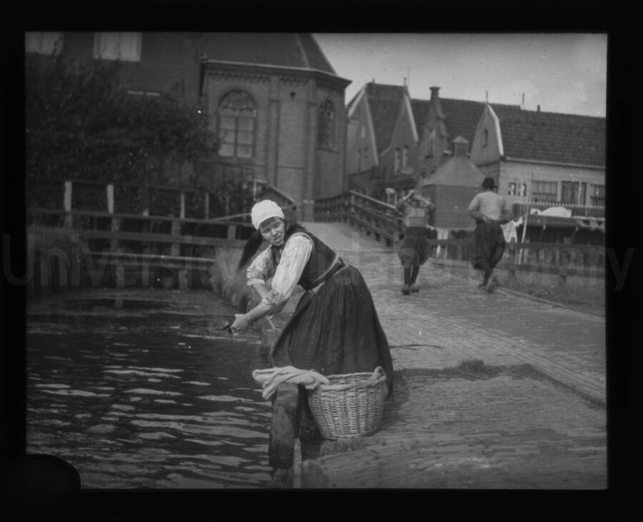 A woman standing by a river alongside a basket filled with clothes on the ground.