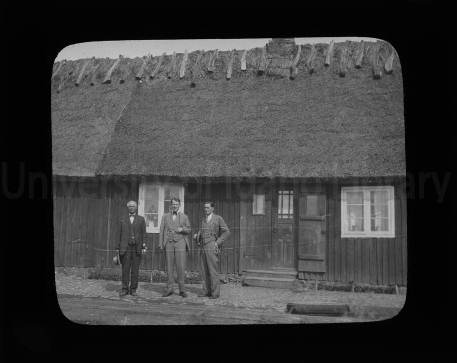 Three men in front of a thatched-roof building.