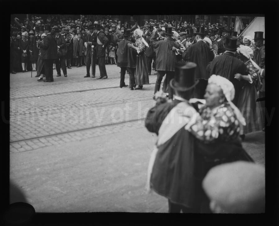 Men in top-hats dance with women in bonnets and flowered dresses during a parade.