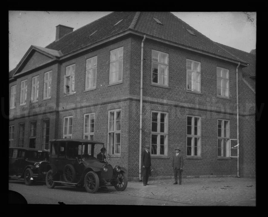 Two cars and three men in front of a building. (Vejen?)