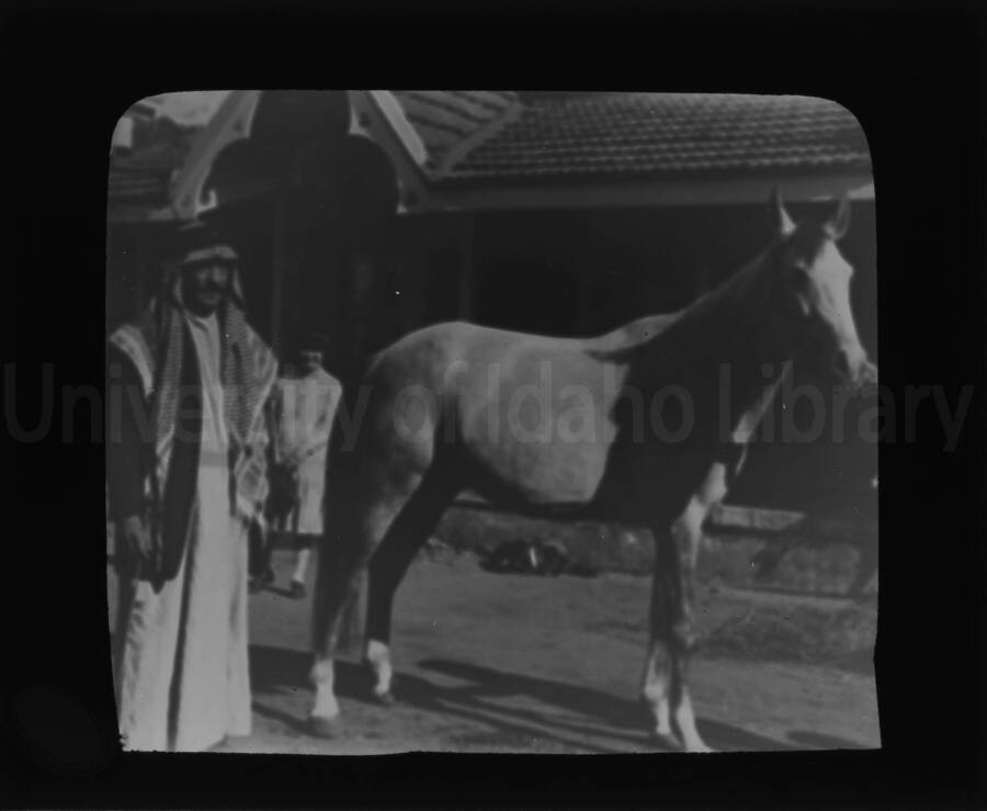 A man in traditional costume with a horse in front of a building in an unknown location.