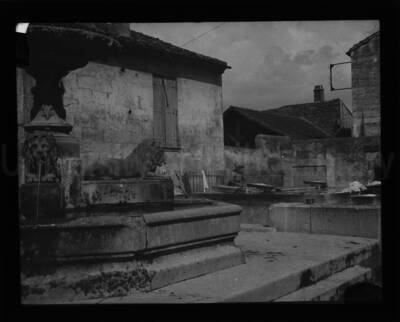 A woman washing laundry in public basins with a fountain in the foreground.