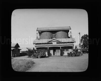 Two cars are parked in front of a building; a brick structure consisting of two joined cylinders stands in the back.