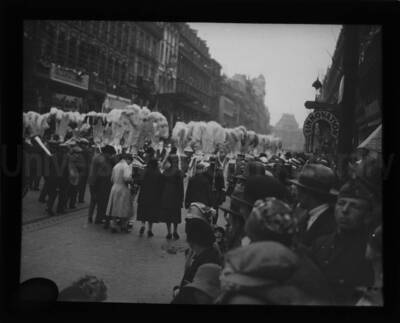 Women follow behind a group of individuals carrying feather topped poles.