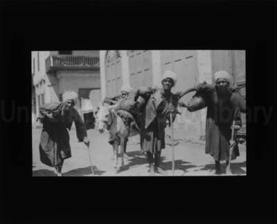 Three men leaning on sticks carrying loaded bags on their shoulders. Behind them what appears to be a man unloading a donkey. (1 duplicate.)