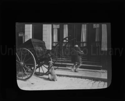 Two-wheeled low trunk carriage parked in front of a hostel. A sign in French on the door of the building advertises rooms for rent. Two girls and a dog are seen on the street.