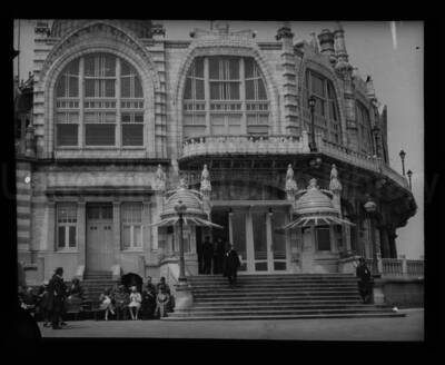 Facade of the Kursaal Oostende, a theater and event space in Oostende, Belgium, destroyed during World War II. Two people sit on benches outside.