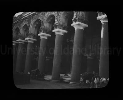 Colonnade of a building with two carriages pulled by horses, in front.