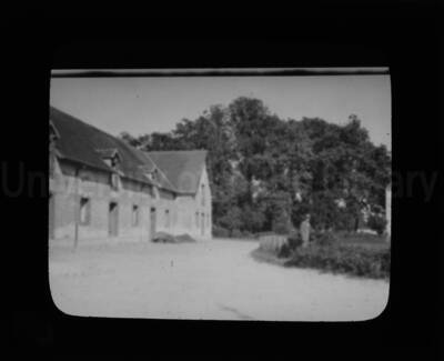 A long building at left and a man standing in a garden, at right.