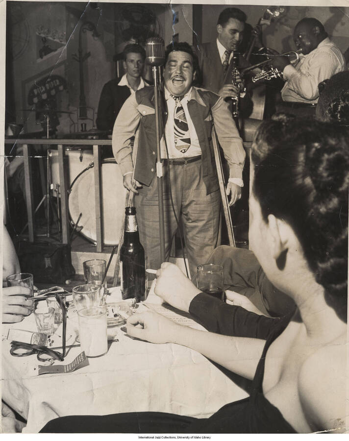 10 x 8 inch photograph; Cornetist Rex Stewart, clarinetist  Buddy DeFranco, and unidentified musicians with audience. The vocalist is using crutches. The background wall includes drawings and the name Pied Piper