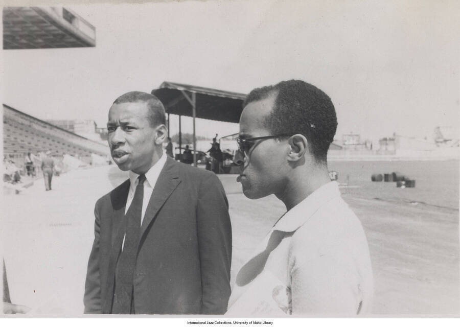 3 1/2 x 5 inch photograph; Trumpeter Lee Morgan is on the left and tenor saxophonist & composer Benny Golson is on the right. They are in a stadium. Observable in the background is an improvised stage with musicians