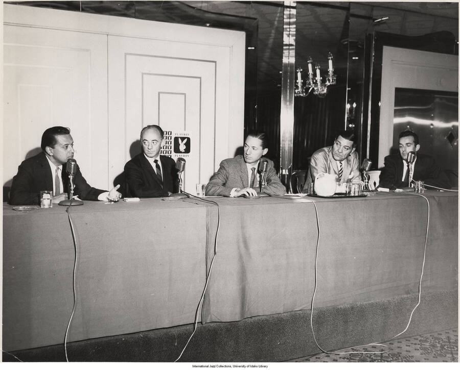 8 x 10 inch photograph; Nesuhi Ertegun, Dave Lambert, Leonard Feather, Jack Teagarden, and Gene Lees at a conference in Chicago. Observable on the wall behind the table is the logo of Playboy Magazine