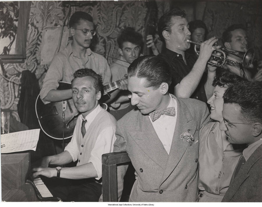 7 x 9 inch photograph; Leonard Feather accompanied by an unidentified man and a woman, observing a pianist. Other musicians in background