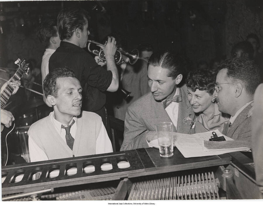 7 x 9 inch photograph; Leonard Feather accompanied by an unidentified man and a woman, observing a pianist. Other musicians in background