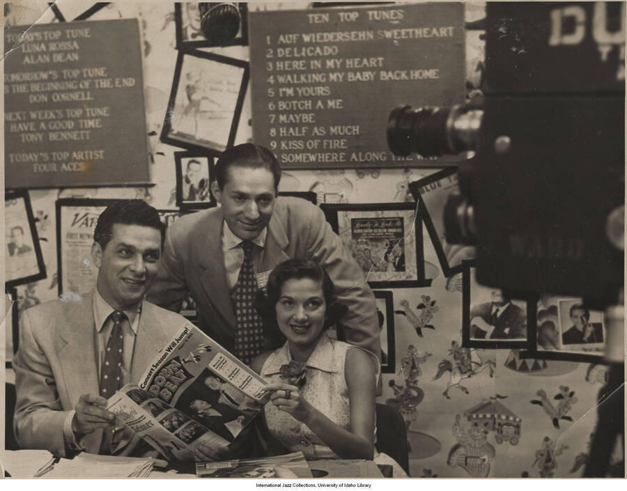 7 x 9 inch photograph; Leonard Feather with unidentified man and woman, in the Du Mont Television studio. They are holding an issue of the Down Beat Magazine from 1952-08