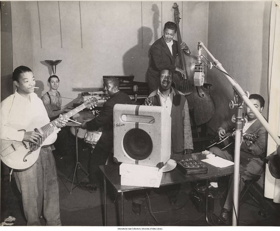 8 x 10 inch photograph; Leonard Feather playing the piano accompanied by Teddy Bunn, on guitar at left singer Leo Watson, George Vann on drums, Red Callender on bass and possibly Ulysses Livingstone on right guitar, in a recording studio.