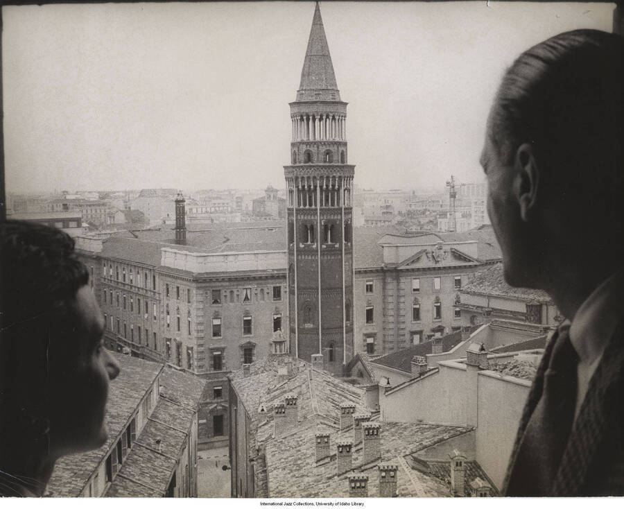 7 3/4 x 9 3/4 inch photograph; Leonard Feather and an unidentified woman looking through a window to the roofs of nearby buildings [in Italy]