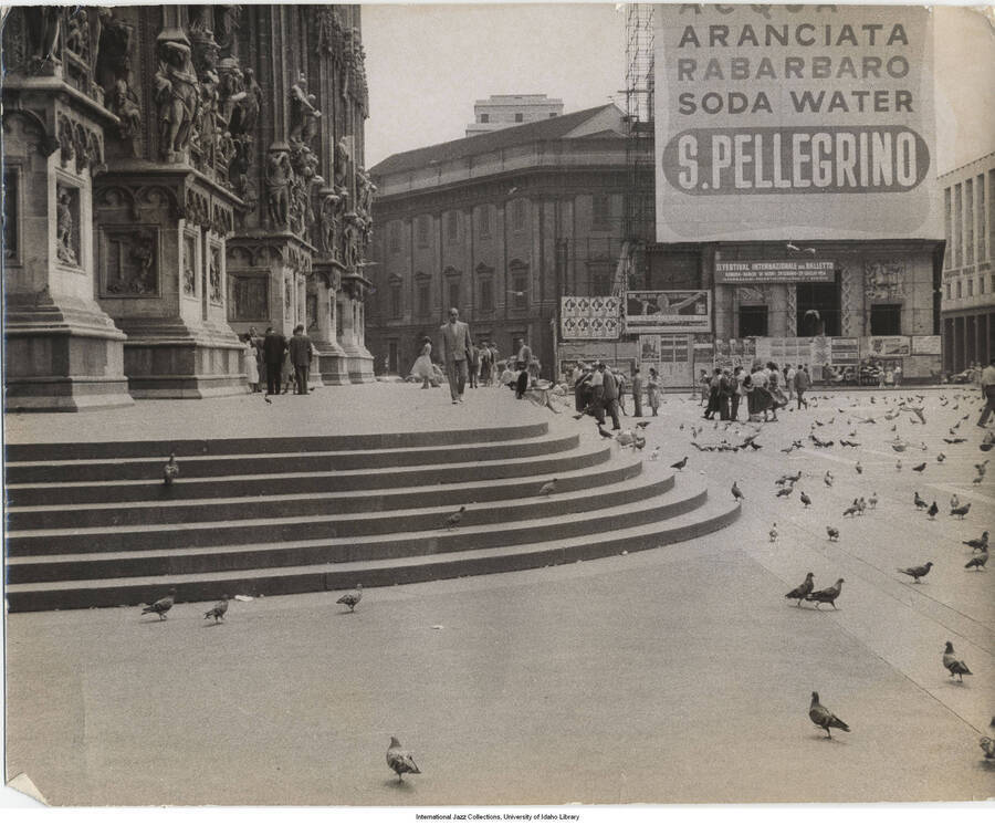 7 3/4 x 9 3/4 inch photograph; sight of a public square surrounded by historical buildings; people and pigeons are seen in the square [in Italy]. Observable in the background is a banner stretched out on the front of a building, probably a theater that reads: II Festival Internazionale del Balletto