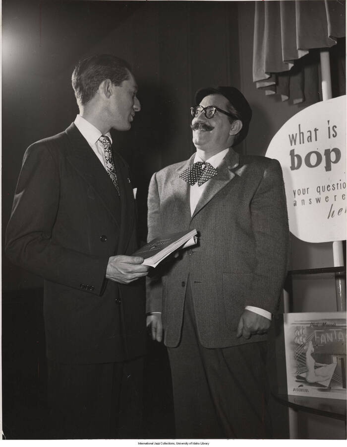 10 x 8 inch photograph. Label on the back of the photograph reads: At Bop City, Leonard Feather (left) offers his new book, "Inside Bebop," to Jerry Colonna as an answer to Jerry's questions about the new jazz. Note Colonna's bop beret, tie, and glasses
