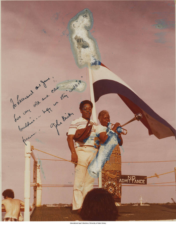 10 x 8 inch signed photograph; Unidentified man and woman standing in front of a flag that includes horizontal stripes from top to bottom, in the colors of red, white and blue. They are standing in what appears to be a boat. The woman is holding a trumpet to her mouth. The photograph is dedicated to Leonard Feather from John Birks.